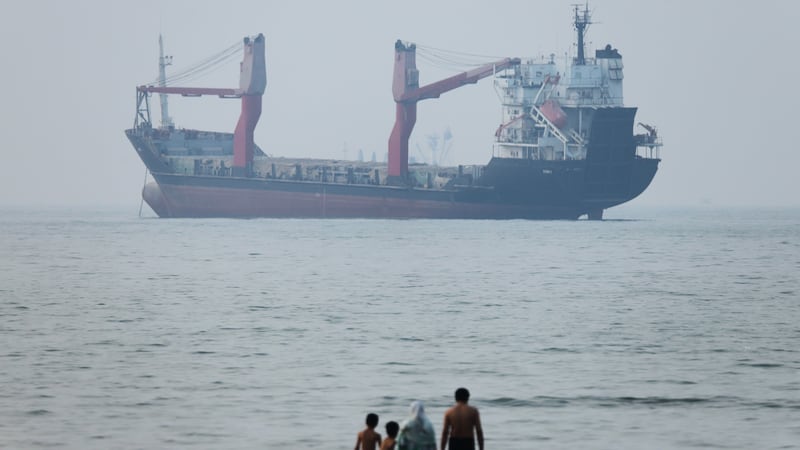 A group of people stands in shallow water as a cargo ship appears anchored in the Strait of...